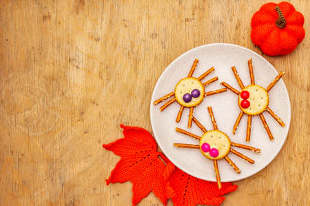 Funny kids food. Edible spiders from cookies, small candies and sweet sesame sticks. Halloween concept with autumn leaves, pumpkin. Ceramic plate, wooden background, copy space, top viewの写真素材