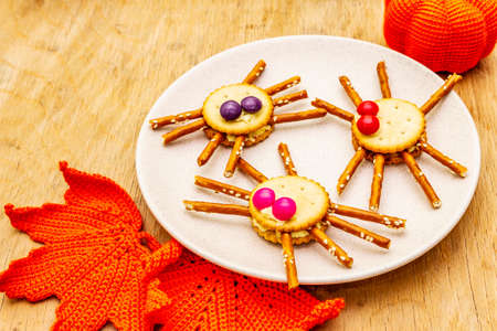 Funny kids food. Edible spiders from cookies, small candies and sweet sesame sticks. Halloween concept with autumn leaves, pumpkin. Ceramic plate, wooden background, close upの写真素材