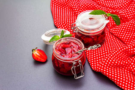 Strawberry jam in a glass jar. Homemade preservation concept. With fresh strawberries and mint, silver spoon on black stone background, polka dots cloth, close up, copy spaceの写真素材