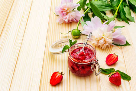 Strawberry jam in a glass jar. Homemade preservation concept. With fresh strawberries and mint, silver spoon, fresh peonies on wooden background, copy spaceの写真素材