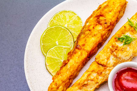 Grilled (fried) pollock in batter. On ceramic plate with lime, parsley and sauce. Cooking fresh fish. On black stone background, close upの写真素材