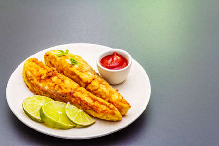 Grilled (fried) pollock in batter. On ceramic plate with lime, parsley and sauce. Cooking fresh fish. On black stone background, copy spaceの写真素材
