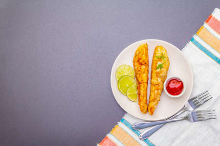 Grilled (fried) pollock in batter. On ceramic plate with lime, parsley and sauce. Cooking fresh fish. On black stone background with vintage linen napkin, steel forks, top view, copy spaceの写真素材