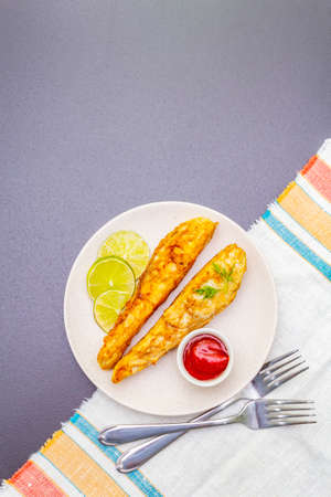 Grilled (fried) pollock in batter. On ceramic plate with lime, parsley and sauce. Cooking fresh fish. On black stone background with vintage linen napkin, steel forks, top view, copy spaceの写真素材