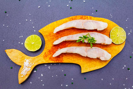 Fresh pollock pieces with lime, green pepper, salt and parsley. Concept of healthy seafood. Organic fish on a wooden board, black stone background, copy space, top viewの写真素材