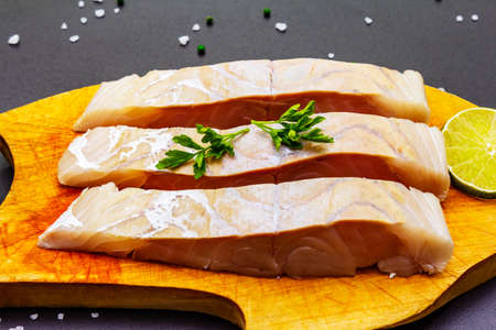 Fresh pollock pieces with lime, green pepper, salt and parsley. Concept of healthy seafood. Organic fish on a wooden board, black stone background, close upの写真素材