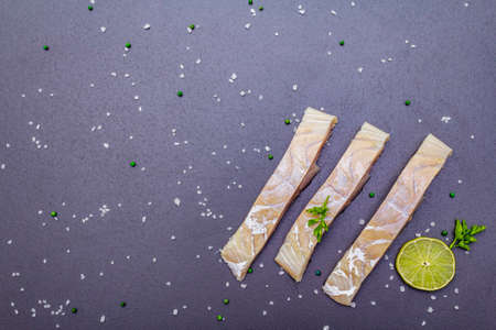 Fresh pollock pieces with lime, green pepper, salt and parsley. Concept of healthy seafood. Organic fish on a black stone background, copy space, top viewの写真素材