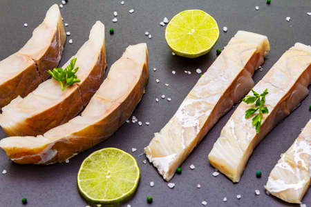 Fresh pollock pieces with lime, green pepper, salt and parsley. Concept of healthy seafood. Organic fish on a black stone backgroundの写真素材