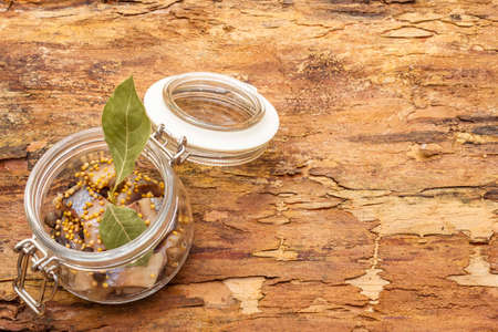 Slices of herring with spices in a glass jar. Mustard seeds, bay leaf, pepper mix. Healthy traditional food concept. Wooden bark background, copy spaceの写真素材