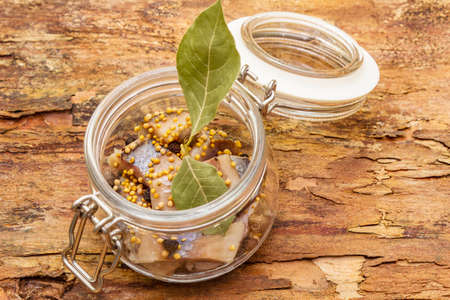Slices of herring with spices in a glass jar. Mustard seeds, bay leaf, pepper mix. Healthy traditional food concept. Wooden bark background, close up, macroの写真素材