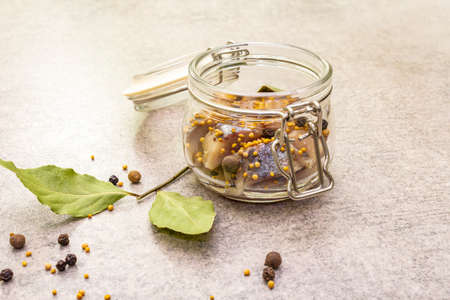 Slices of herring with spices in a glass jar. Mustard seeds, bay leaf, pepper mix. Healthy traditional food concept. Stone background, close upの写真素材