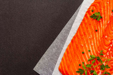 A piece of raw trout fillet (salmon) with parsley, salt and green pepper. Ingredient for cooking healthy food. Fresh fish on a parchment, black matte background, copy space, top view, close upの写真素材