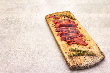 Basturma, dried tenderloin of beef meat, jerky, thinly sliced. Dry rosemary, pepper mix on vintage wooden board. Delicious food on a stone background, copy spaceの写真素材