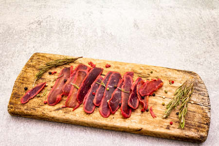Basturma, dried tenderloin of beef meat, jerky, thinly sliced. Dry rosemary, pepper mix on vintage wooden board. Delicious food on a stone backgroundの写真素材