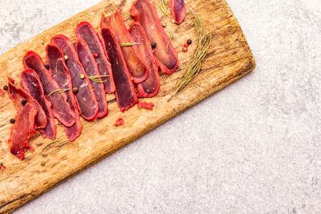 Basturma, dried tenderloin of beef meat, jerky, thinly sliced. Dry rosemary, pepper mix on vintage wooden board. Delicious food on a stone background, top view, copy space, close upの写真素材