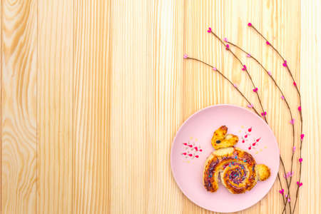 Fresh bun in the form of an Easter bunny. The concept of children's holiday bakery. In pink ceramic plate with artificial willow branches on wooden background, copy space, top viewの写真素材