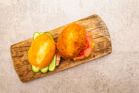 Homemade sandwich in bun. Salami, dried pork, tomato, cucumber, cheese ingredients. On wooden board and stone background, top viewの写真素材