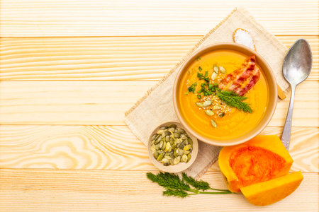 Homemade autumn hot pumpkin cream soup with smocked bacon and seeds. Raw pumpkin, fresh chives, dill, salt in spoon, vintage linen cloth on wooden background, top view, copy spaceの写真素材