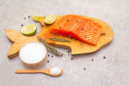 Raw trout (salmon) fillet piece with lime, rosemary, sea salt and pepper mix. Wooden board in the shape of a fish, on a stone background.の写真素材