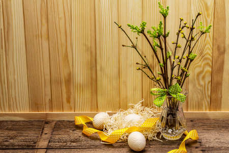Zero waste Easter concept. Spring twigs with fresh green leaves. Glass vase, wooden eggs, nest, polka dot ribbon. Wooden boards background and backdrop, copy spaceの写真素材