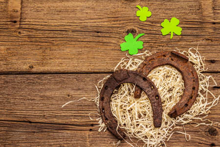 Two cast iron metal horse horseshoes with felt clove leaves on hay. Good luck symbol, St.Patrick's Day concept. Old wooden background, horse accessories, top viewの写真素材