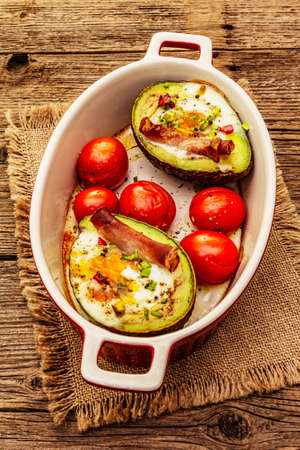 Sunny morning breakfast. Baked eggs in avocado half, bacon, tomato cherry, spices. Healthy food, ketogenic diet concept. Old wooden boards backgroundの写真素材