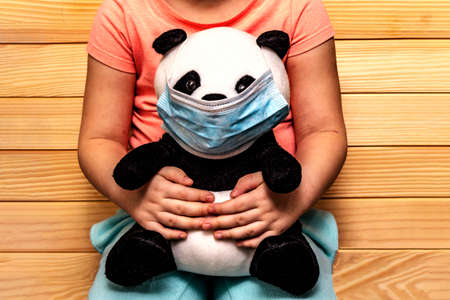 Soft toy panda in a disposable medical mask in children's hands. Coronavirus epidemic concept. Personal hygiene for kids. Wooden backgroundの写真素材