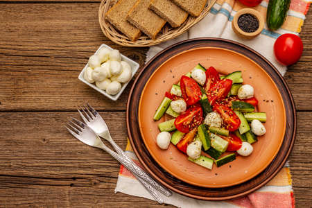Fresh salad from tomato, cucumber and baby mozzarella. Sunny lunch with crusty bread. Old wooden boards background, top viewの写真素材