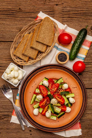 Fresh salad from tomato, cucumber and baby mozzarella. Sunny lunch with crusty bread. Old wooden boards background, top viewの写真素材