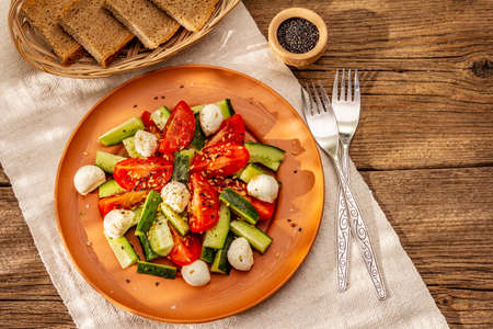 Fresh salad from tomato, cucumber and baby mozzarella. Sunny lunch with crusty bread. Old wooden boards background, top viewの写真素材