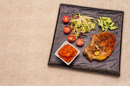 Grilled pork steak. Cole slow salad, tomato sauce, fresh vegetables. Beige table background, top viewの写真素材