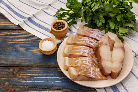 Raw fillet of saithe. Fresh slices on a plate, spices, parsley. A bunch of greens, vintage napkin. Old wooden boards background, copy spaceの写真素材