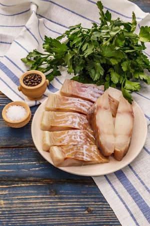 Raw fillet of saithe. Fresh slices on a plate, spices, parsley. A bunch of greens, vintage napkin. Old wooden boards background, copy spaceの写真素材