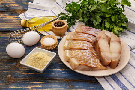 Raw fillet of saithe and frying ingredients. Fresh slices on a plate, spices, eggs, oil, breadcrumbs, parsley. A bunch of greens, vintage napkin. Old wooden boards background, close upの写真素材