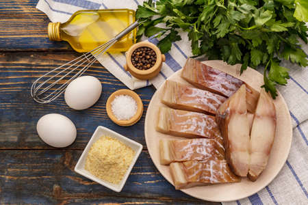Raw fillet of saithe and frying ingredients. Fresh slices on a plate, spices, eggs, oil, breadcrumbs, parsley. A bunch of greens, vintage napkin. Old wooden boards background, top viewの写真素材