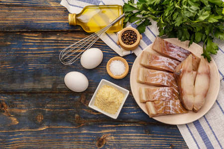 Raw fillet of saithe and frying ingredients. Fresh slices on a plate, spices, eggs, oil, breadcrumbs, parsley. A bunch of greens, vintage napkin. Old wooden boards background, top viewの写真素材