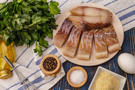 Raw fillet of saithe and frying ingredients. Fresh slices on a plate, spices, eggs, oil, breadcrumbs, parsley. A bunch of greens, vintage napkin. Old wooden boards background, close upの写真素材