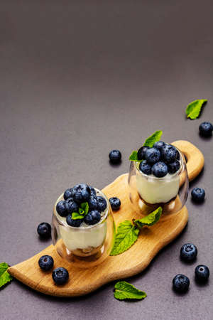 Yogurt with blueberries and chia seeds on wooden cutting board. Healthy and useful breakfast concept. Ripe berries, mint leaves, black stone concrete background, copy spaceの写真素材