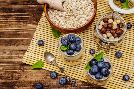 Healthy and useful breakfast concept. Yogurt with blueberries, oat-flakes and chia seeds. Ripe berries, mint leaves, silver spoon on wooden boards backgroundの写真素材