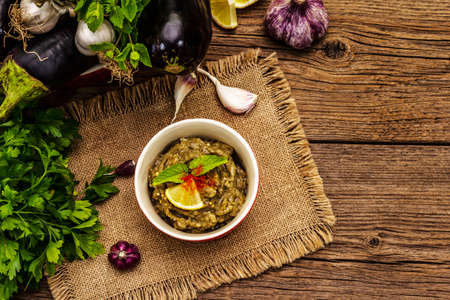 Eggplant dip baba ganoush (mutabbal) or mezze with ripe vegetables and fresh herbs. Traditional Middle East cuisine, healthy appetizer, vegan hummus. Old wooden boards background, top viewの写真素材