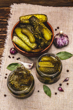 Marinated pickled cucumbers with spices and herbs. Traditional fermented appetizer in glass jars and clay bowl. Wooden boards background, top viewの写真素材