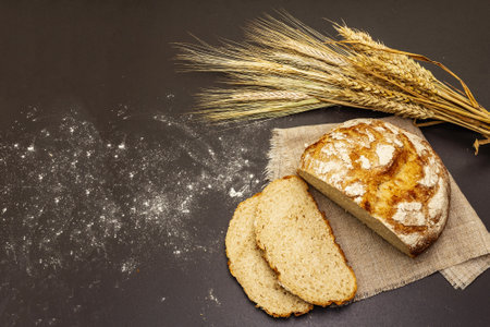Fresh baked bread with wheat ears on wooden stand. Slices and linen napkin. Black stone concrete...