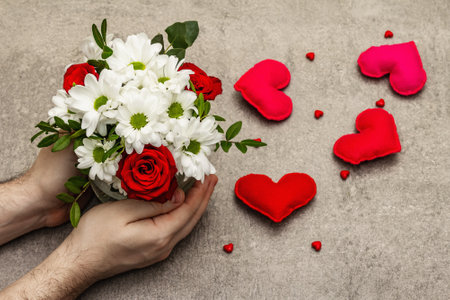 Valentine's Day or Happy Birthday concept. Male hands are holding a bouquet of flowers. Scattered handmade felt hearts, gray stone concrete backgroundの写真素材
