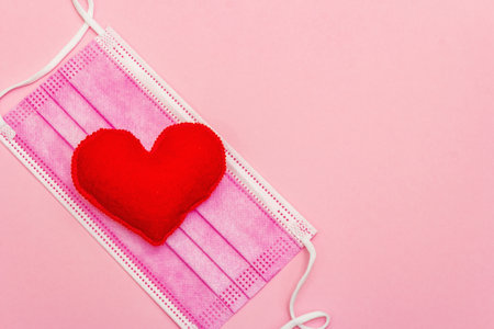 Red heart with medical protection mask on light pink background, top view, copy space. Concept of...