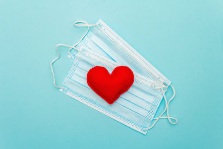 Red heart with medical protection masks on light blue background, top view, copy space. Concept of...