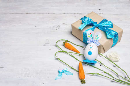 Handmade traditional Easter symbols concept. Cute rabbit from the egg, gift box, festive decor in blue tones. White wooden background, copy spaceの写真素材