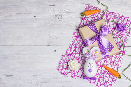 Handmade traditional Easter symbols concept. Cute rabbit from the egg, gift box, festive decor in lilac tones. White wooden background, top viewの写真素材