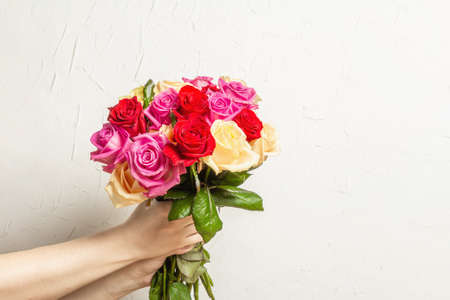 The woman's hands are holding a beautiful bouquet of fresh multicolored roses. Flower composition on white plaster background. Festive card conceptの写真素材