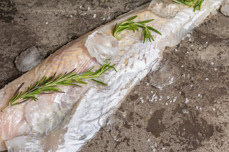 Raw frozen hake fillet with ice and fresh rosemary. Healthy food concept, the cooking fish process. Dark marble stone background, top viewの写真素材