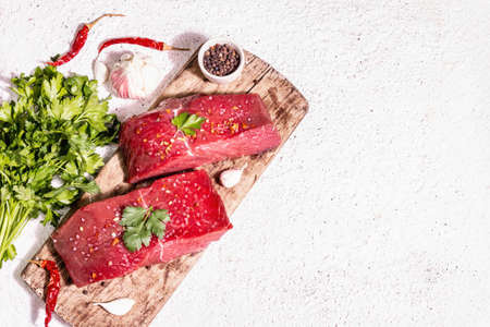 Raw beef steaks on a wooden cutting board. Trendy hard light, dark shadow. Spices, hot chili, fresh garlic, and parsley. Light plaster background, top viewの写真素材